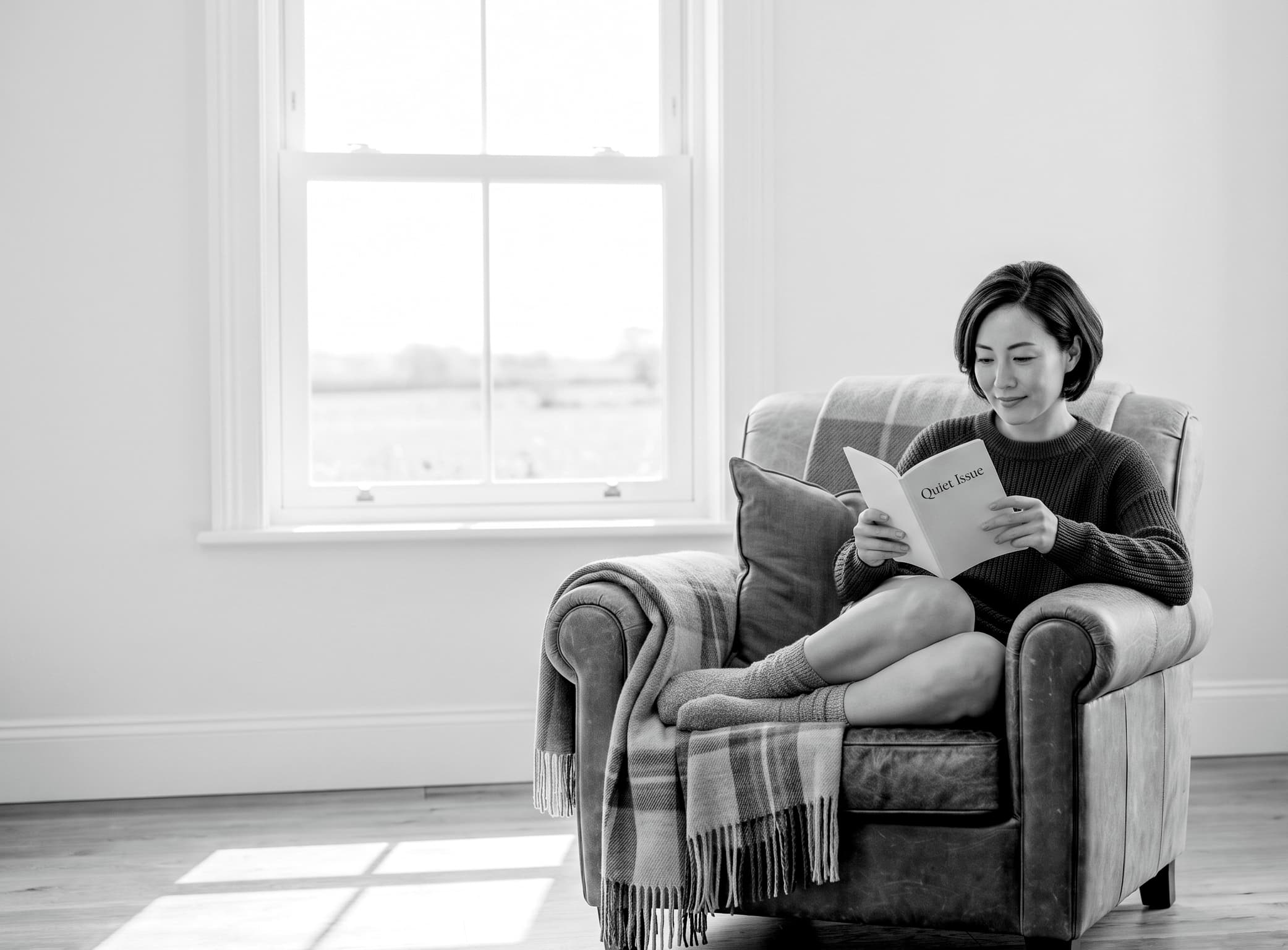 A woman reading a Quiet Issue booklet in a leather armchair by a sunlit window